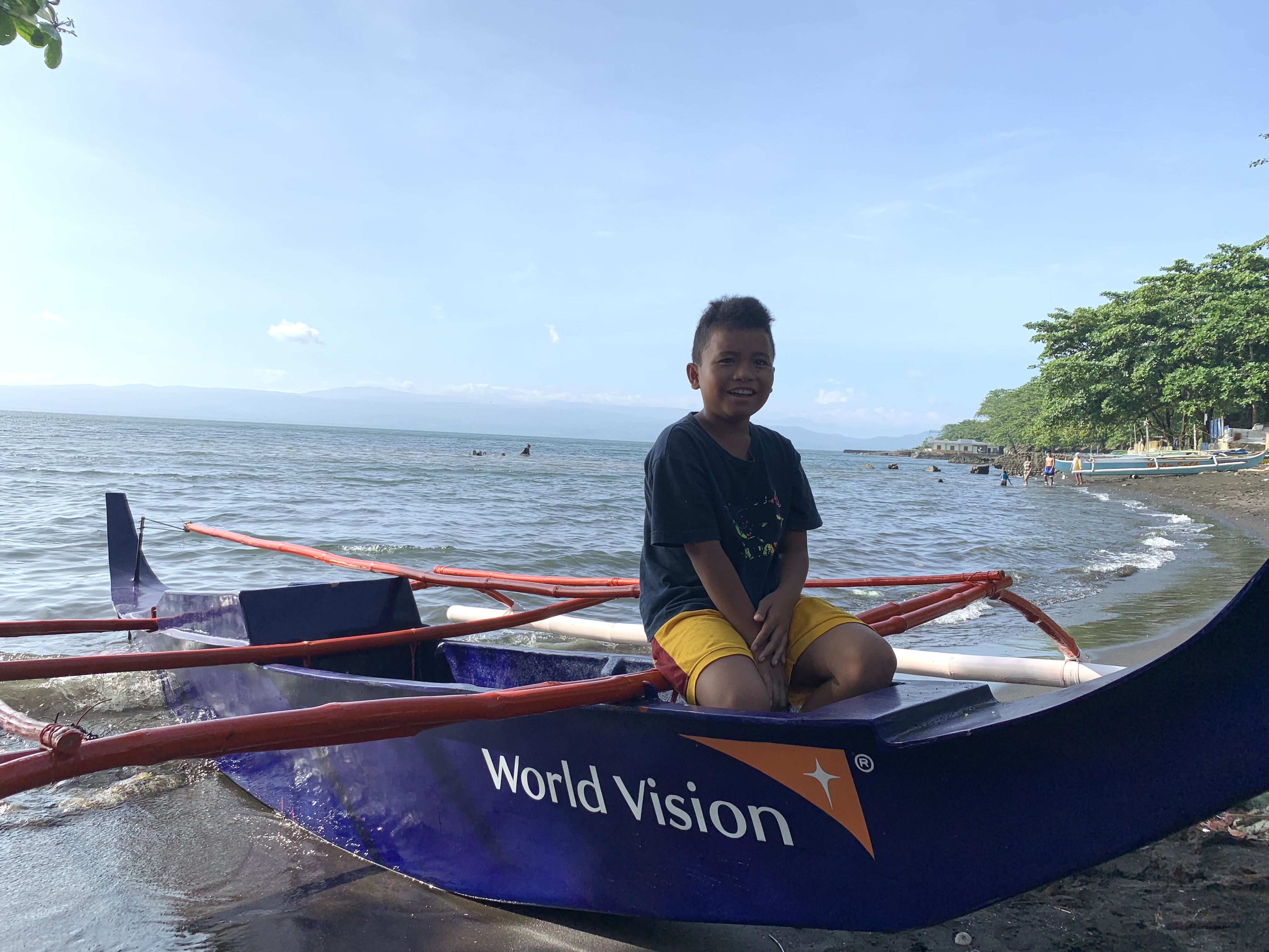 11-year-old boy perches on the side of a blue wooden fishing boat, on the shore. Sea and clear blue sky behind