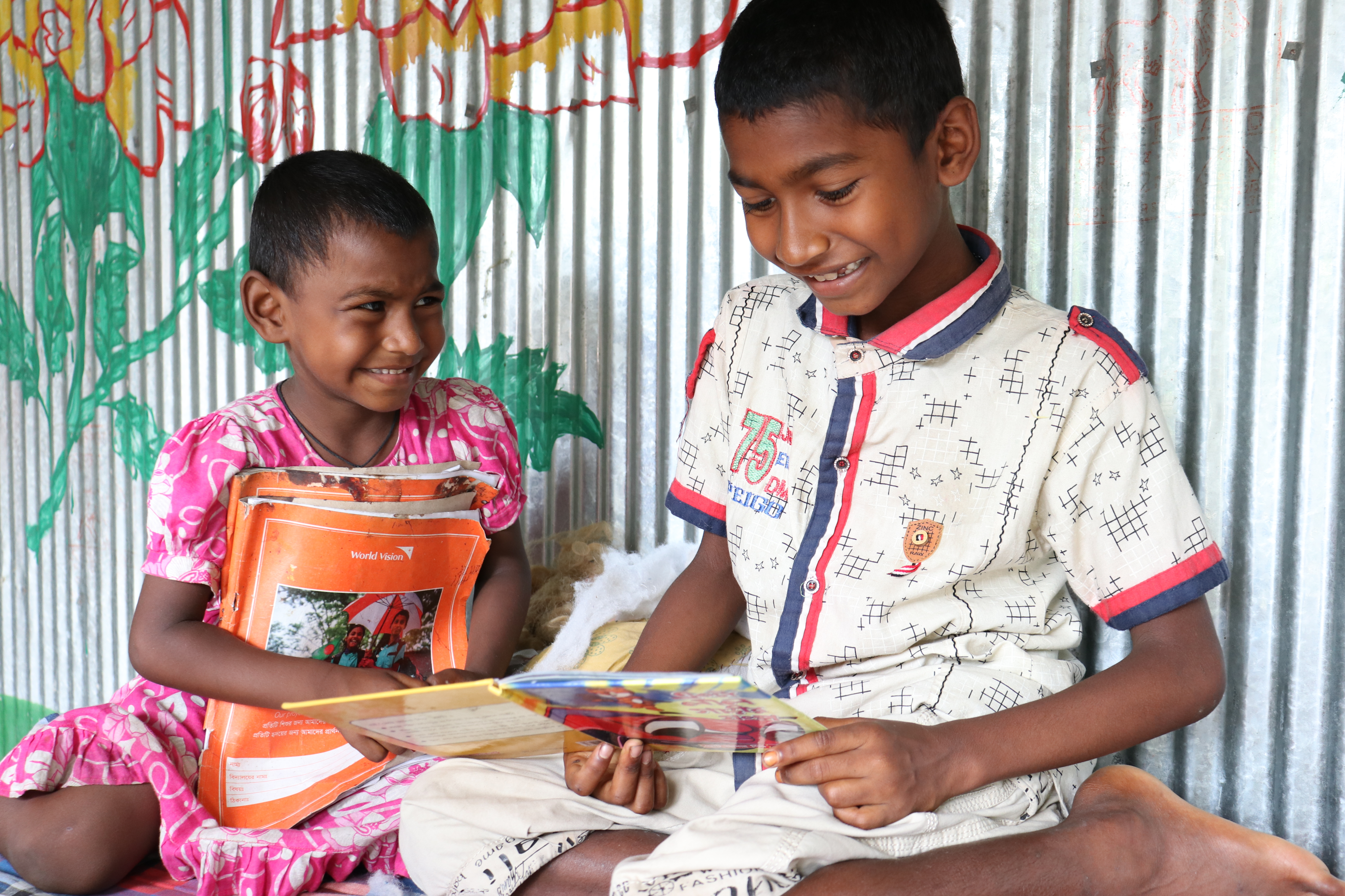 Two children from Bangladesh sat looking at educational activity pack and smiling