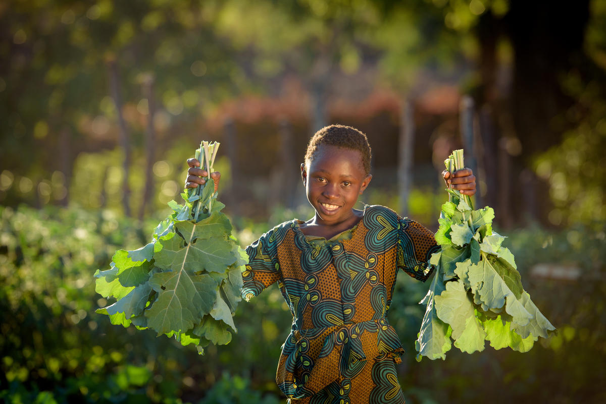 A young girl in Zambia in a traditional green dress holds leafy vegetables up and smiles at the camera