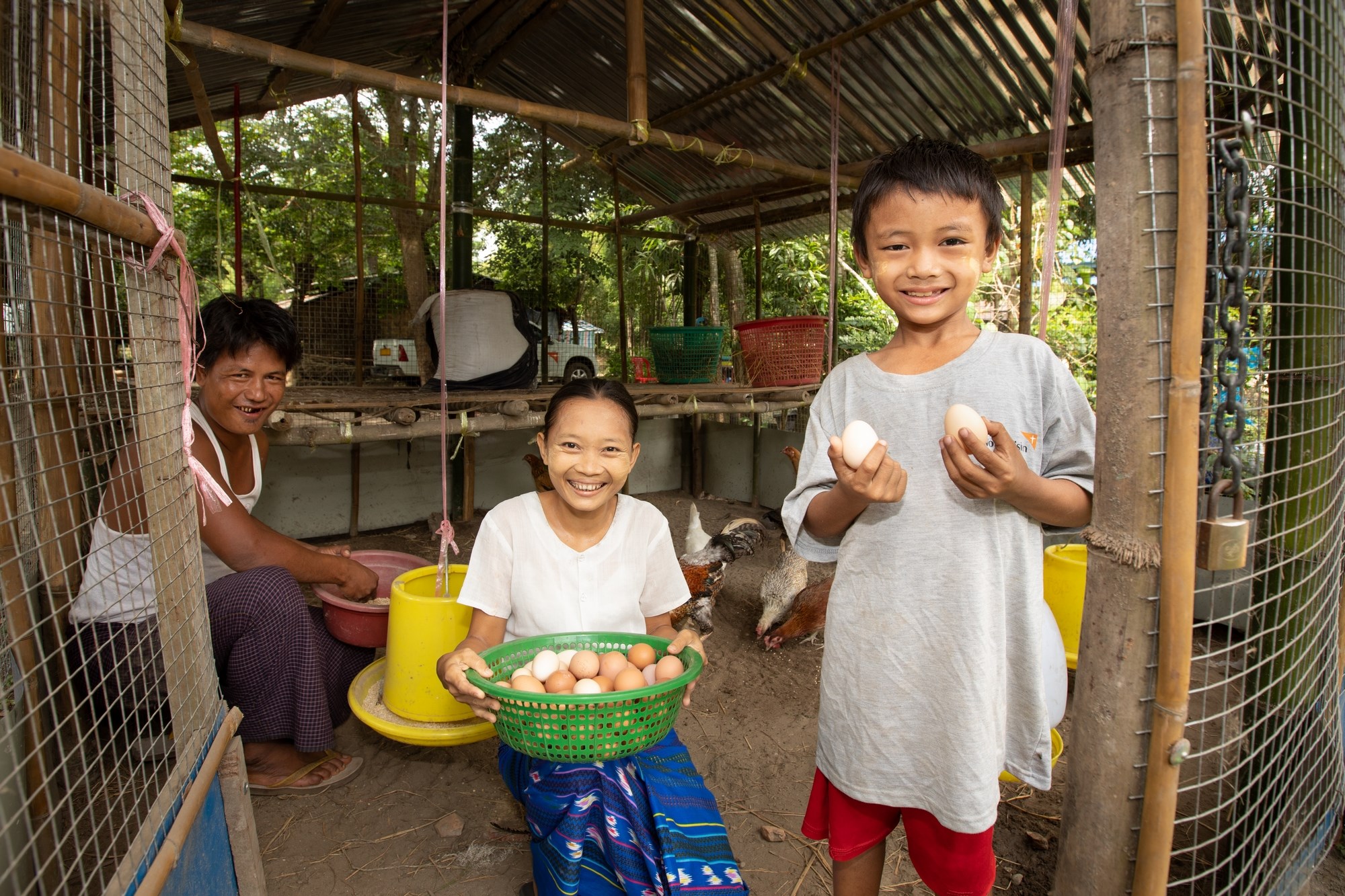 A young girl holding a bowl of eggs, and a boy holding an egg in each hand. Standing in front of their family smallholding in Myanmar