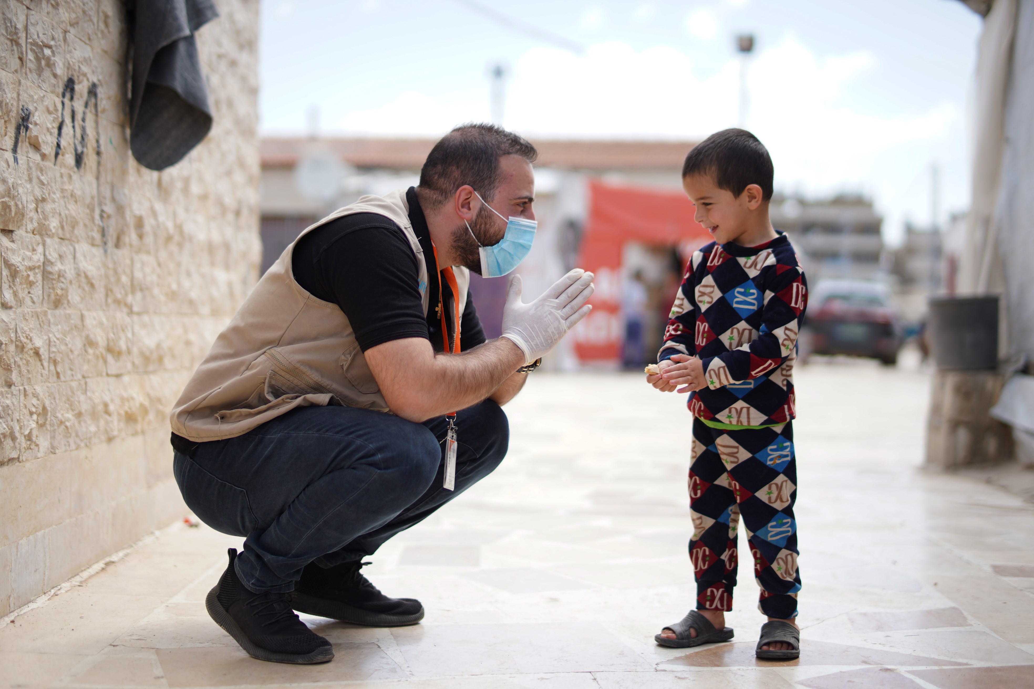 A World Vision member in a refugee camp in Lebanon wearing a mask kneels down to talk to a boy wearing pajamas