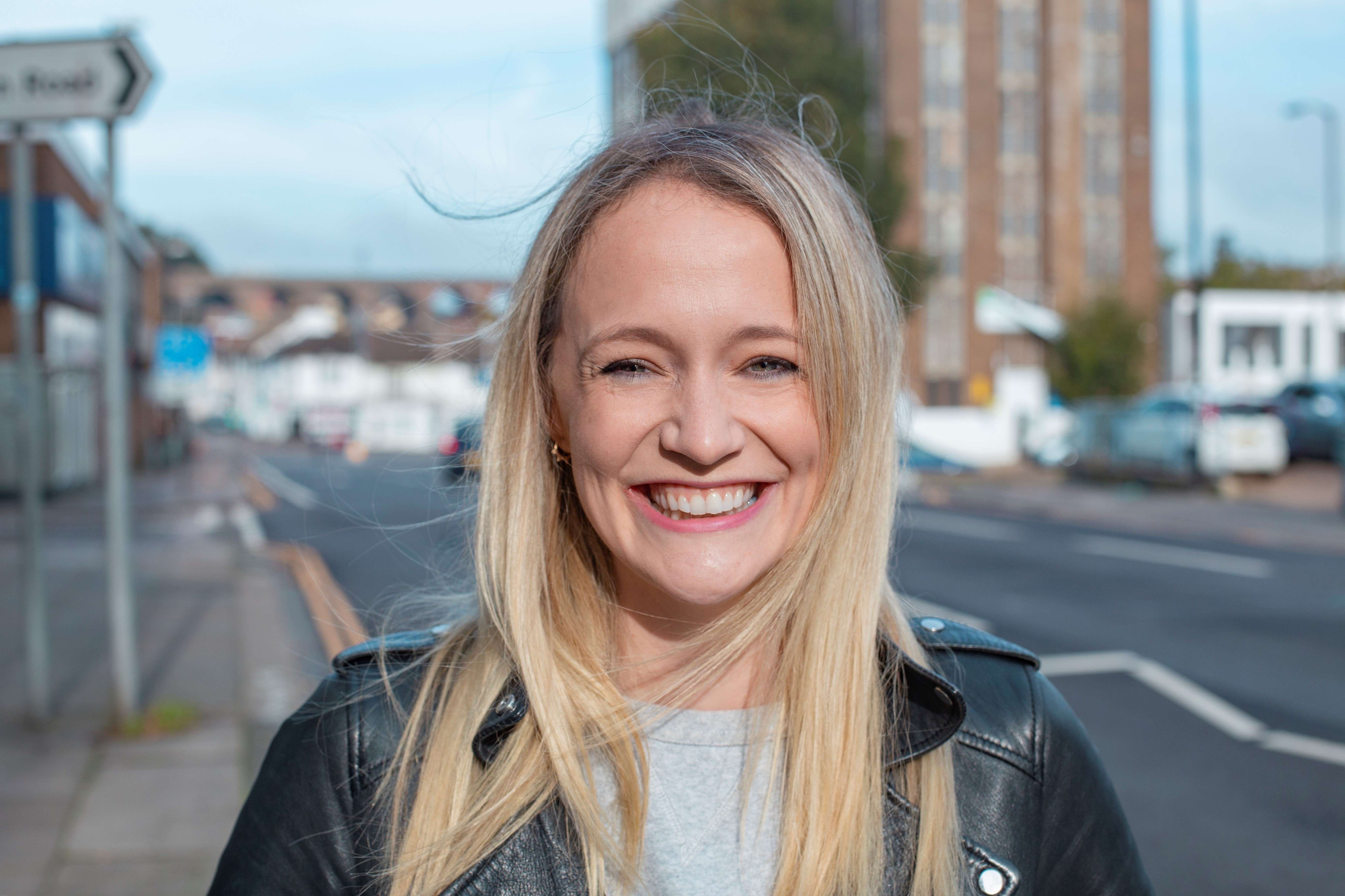 Woman in the UK smiles broadly, standing on a street against a blue sky