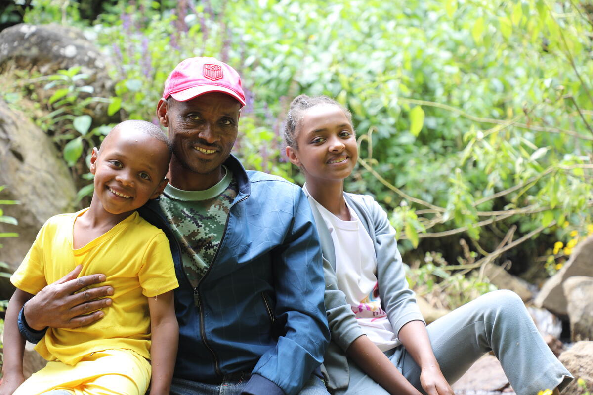 A father poses with his son and daughter who are leaning on his sides. There's a farm behind them.