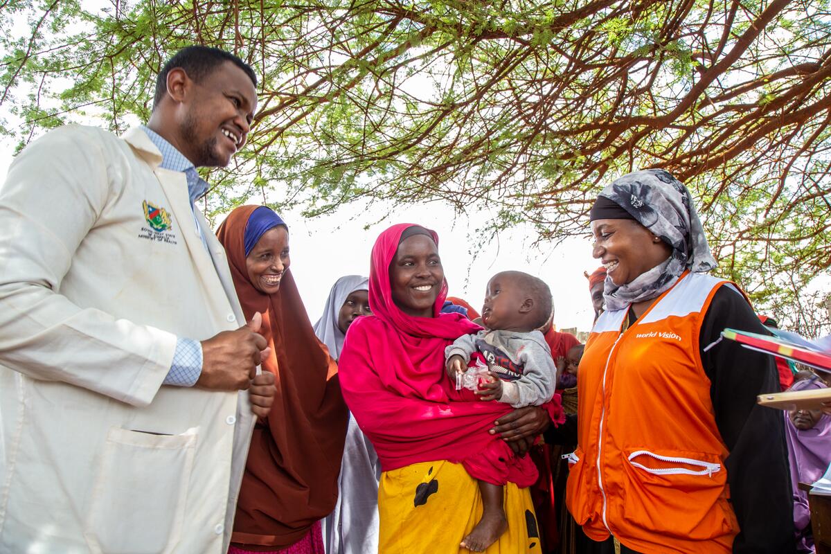 Female doctor holds out an injection over a desk, leaning towards a mother. The child on her lap looks nervously at the syringe