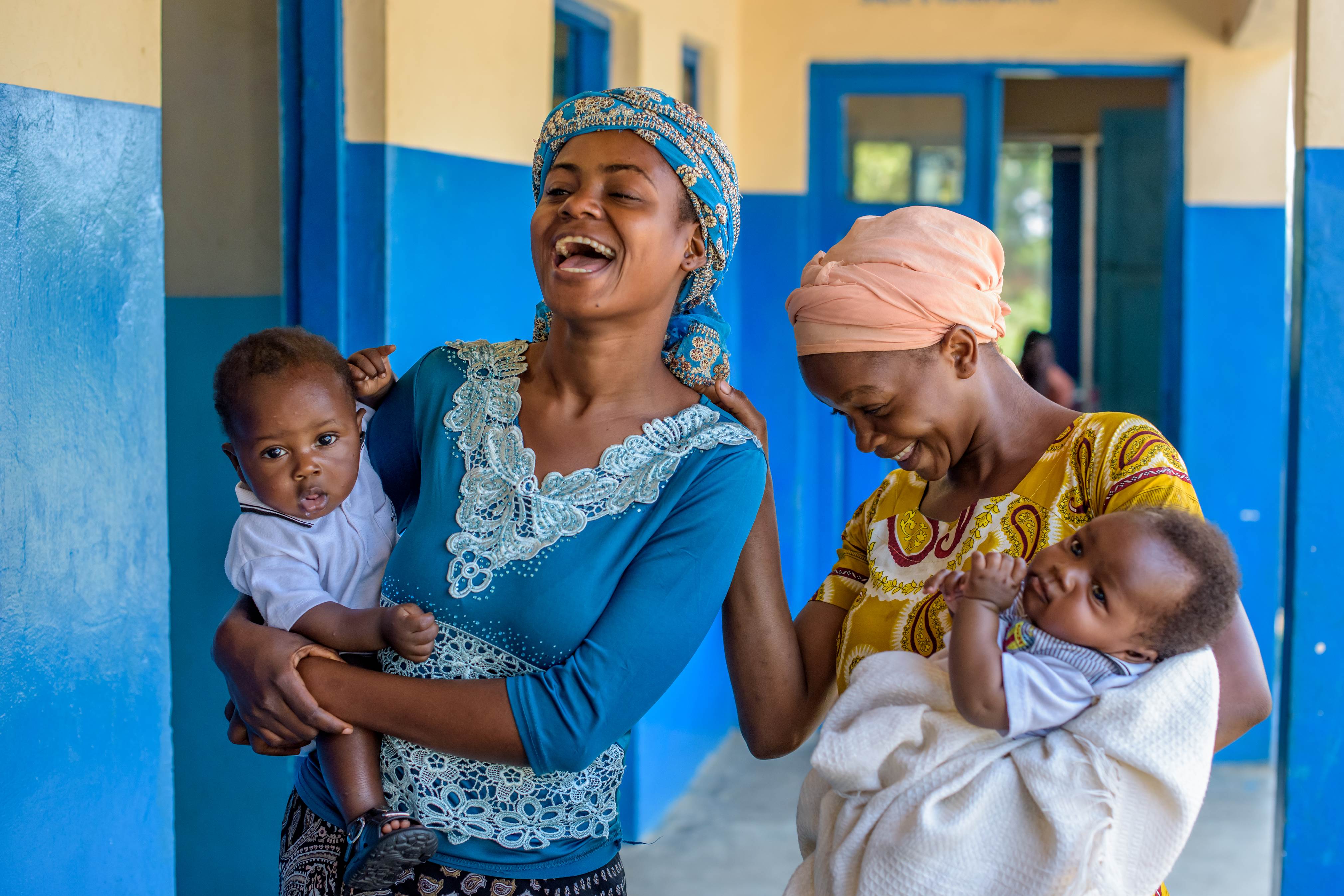 Two mothers laughing with each other while each is holding a baby in their arms with building walls behind them
