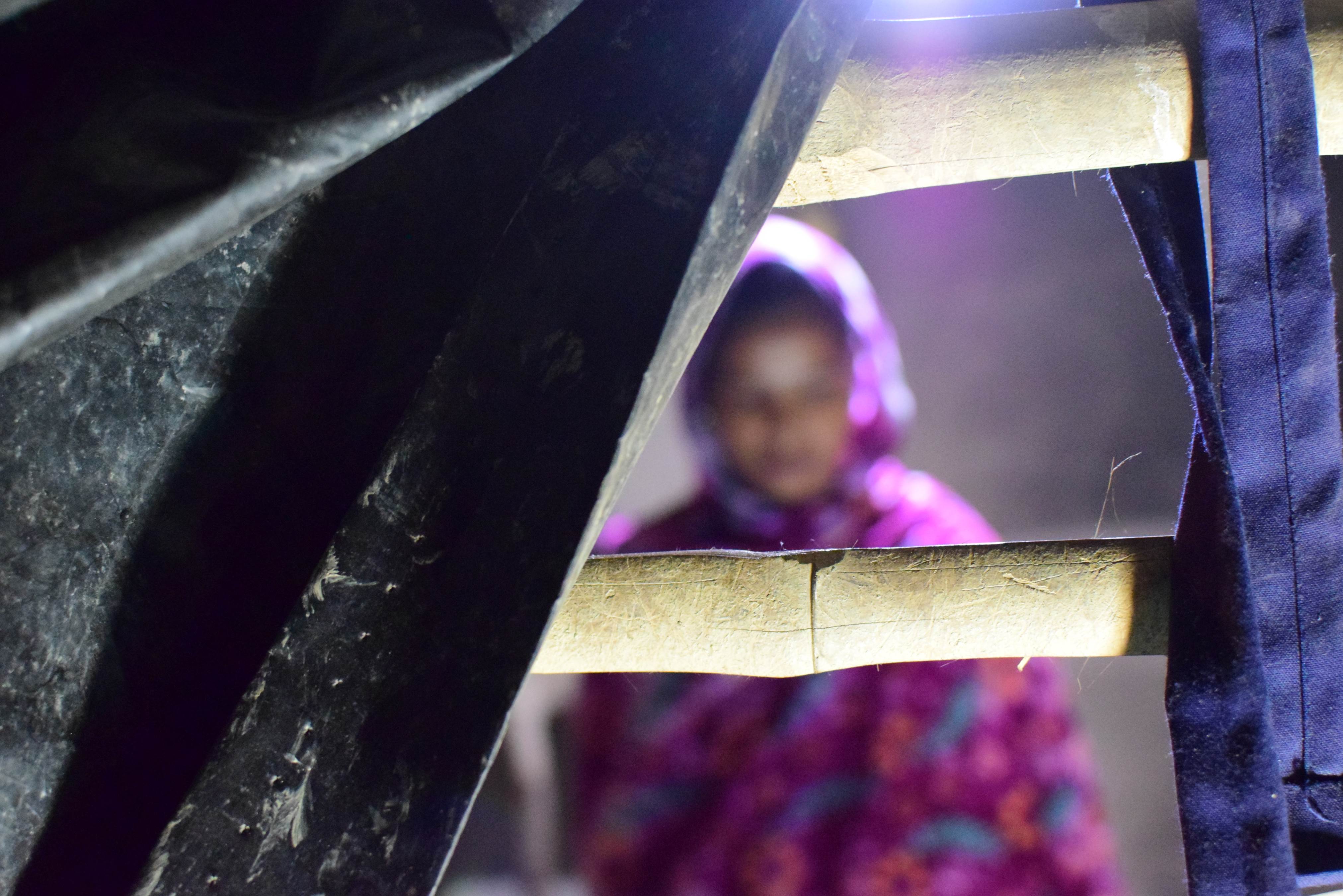 Girl from India wearing a Hijab, sat in a dimly lit room. The photo is taken from behind a barrier of bamboo sticks