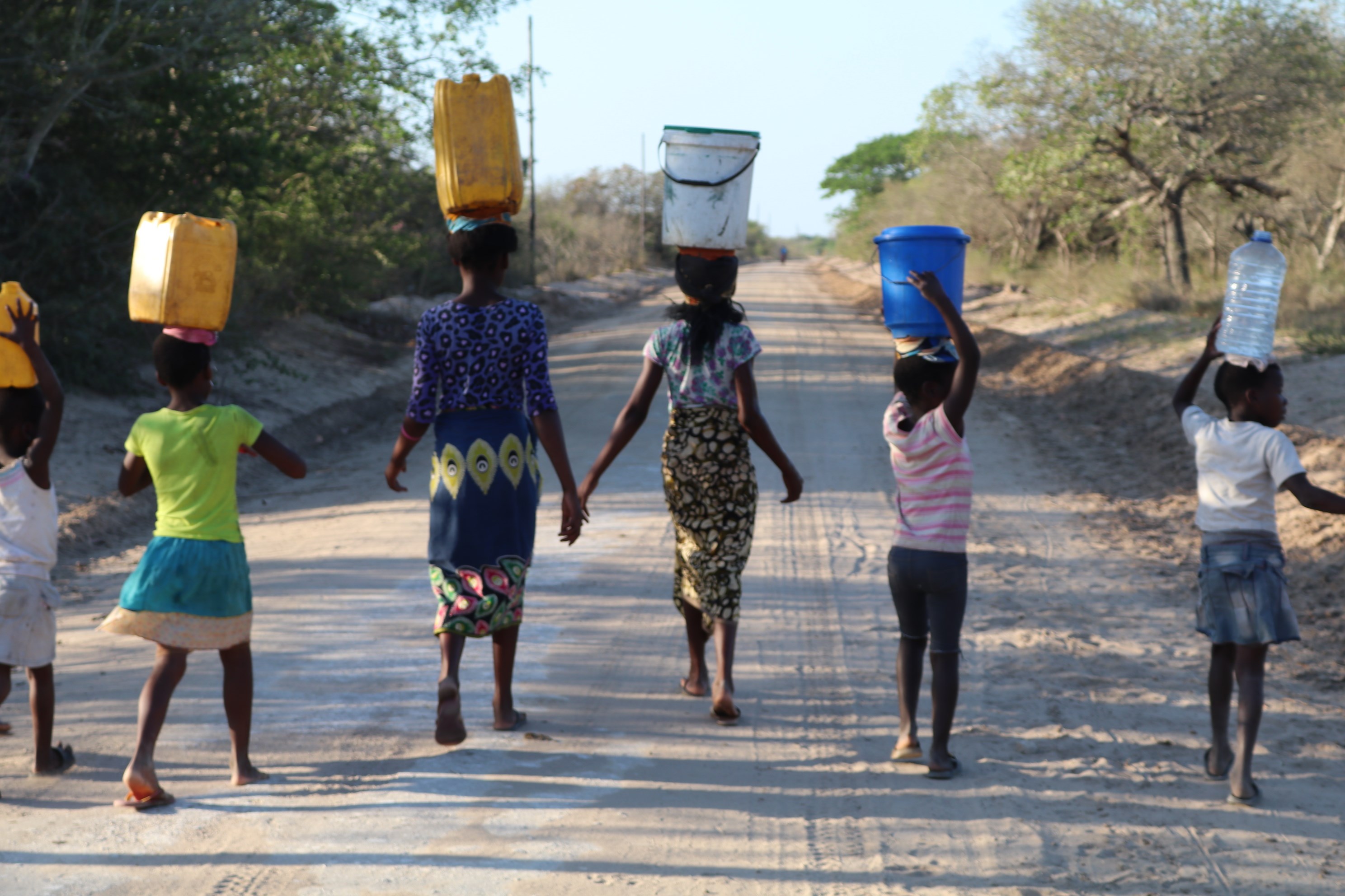 Group of people from Mozambique walk away from camera along a road, holding water bottles on their head
