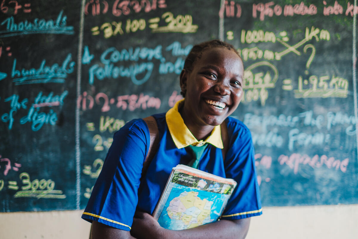 A girl in school uniform, stood in front of a blackboard, smiling and holding a book in her arms 
