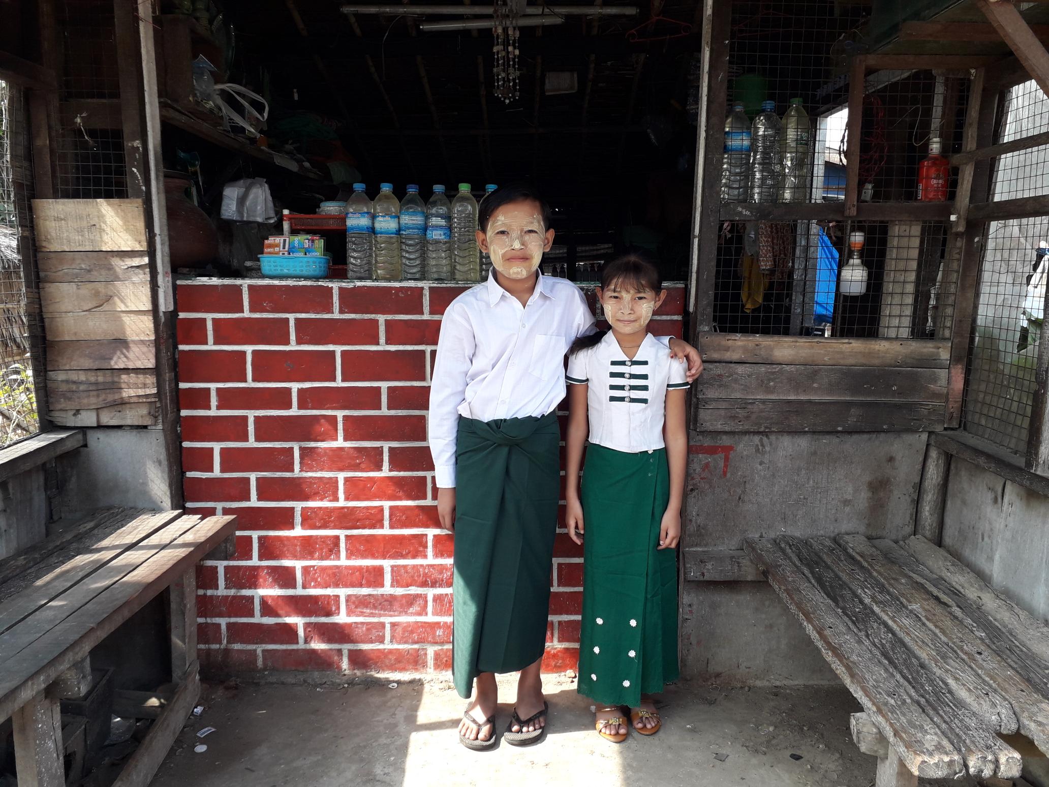 A girl and boy from Myanmar wearing school uniforms stand together in front of their school's entrance