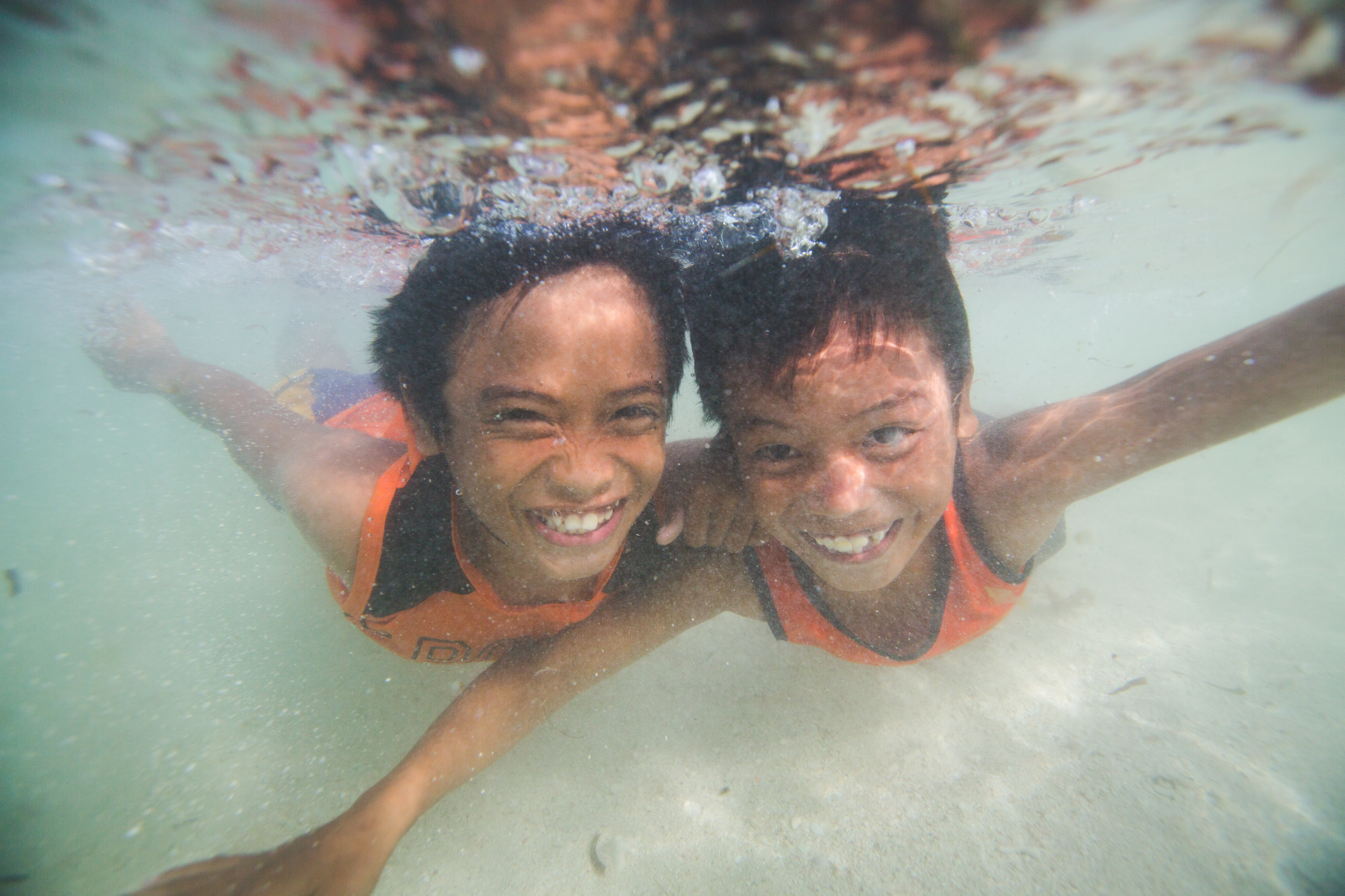 Two young boys in orange vests, swim underwater towards the camera