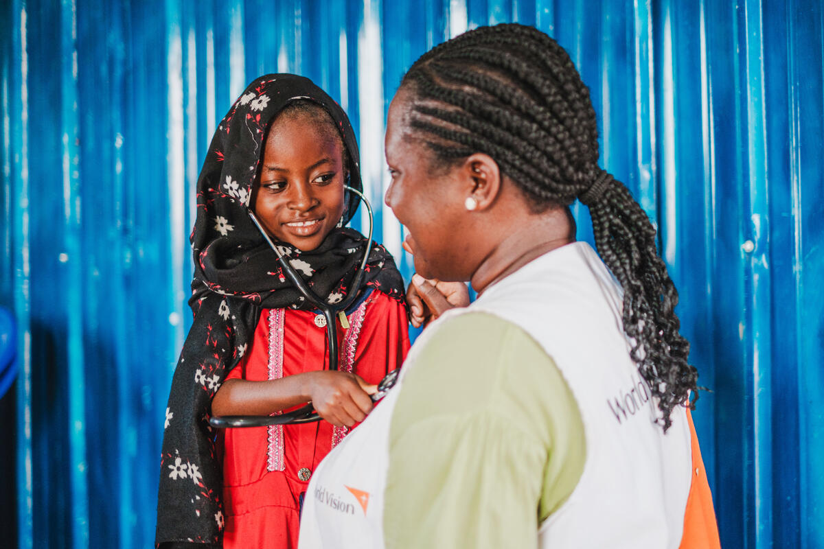 A young girl holding a stethoscope to a World Vision staff member and is smiling.