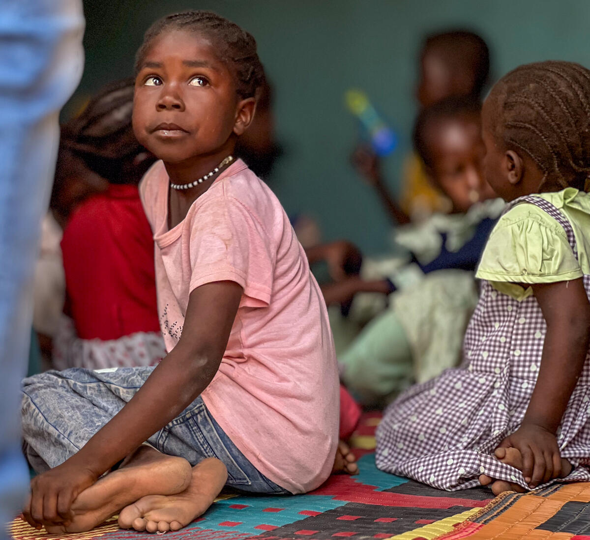 A young girl among a group sat on the floor turns sideways and looks away from the camera