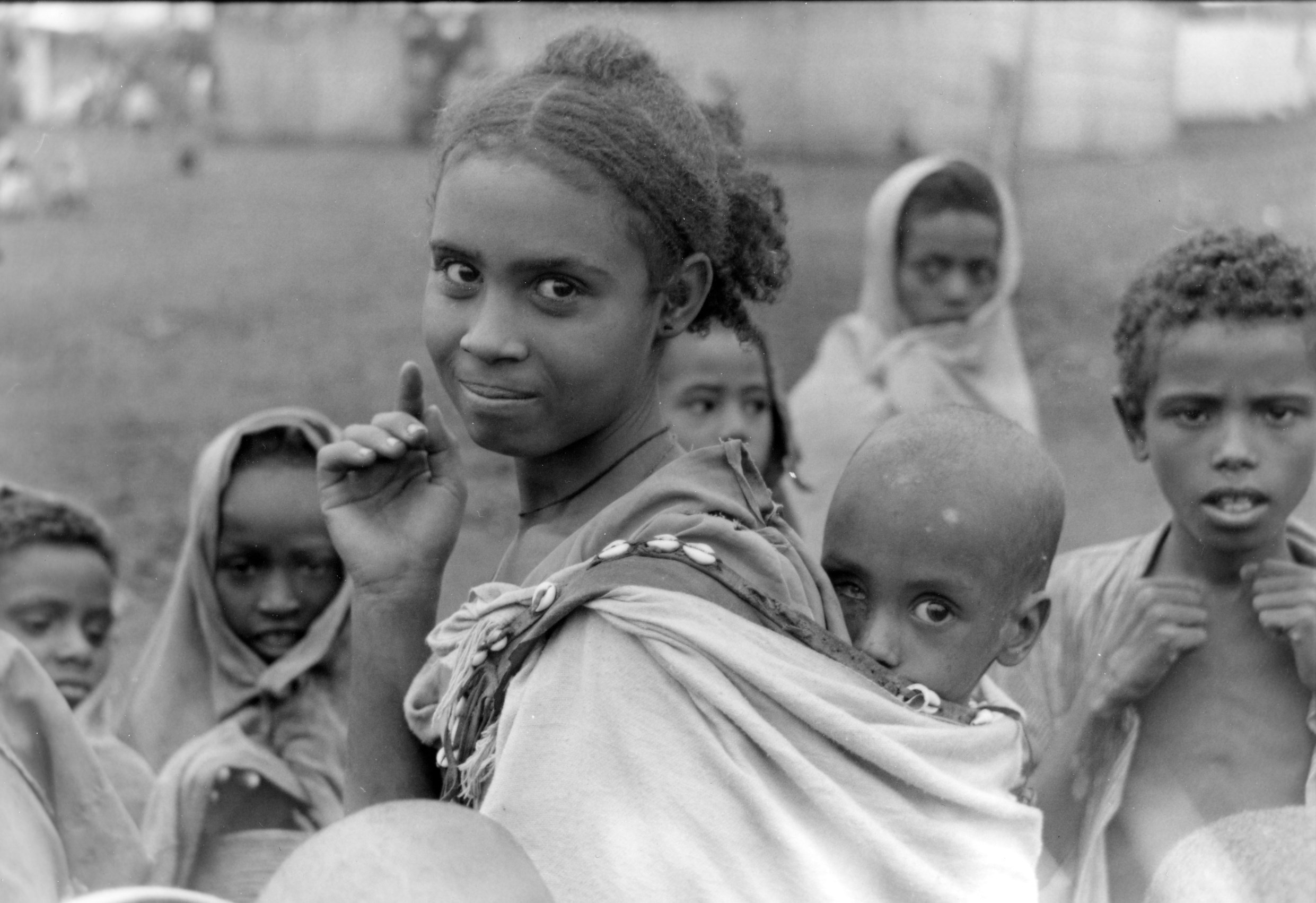 Woman looks into camera with baby in a sling on her back, and other children behind her also looking at the camera in this black and white photo
