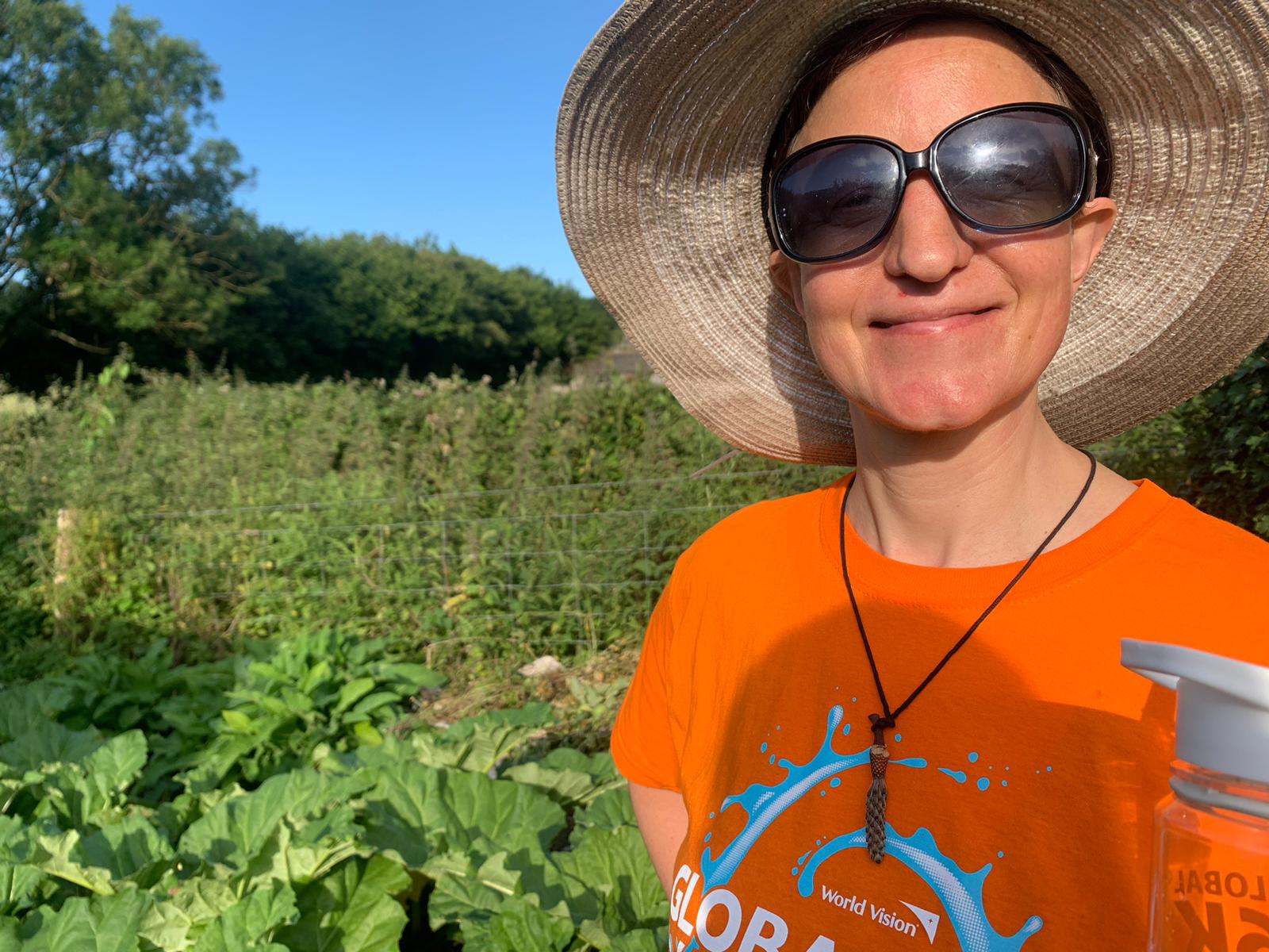 A woman poses for a photo in her garden. She has a straw hat and sunglasses on.