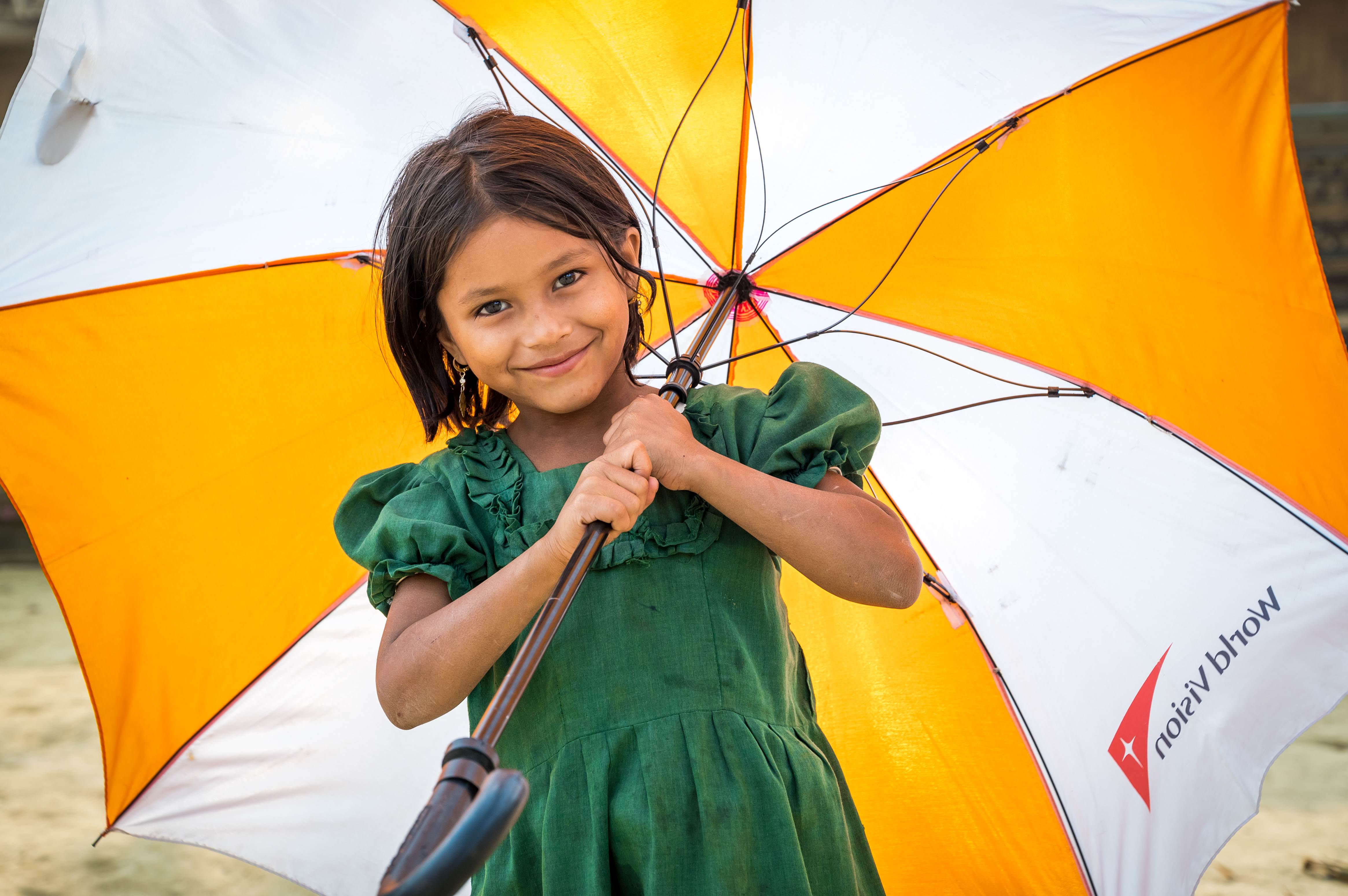 A young Rohingya refugee, smiles as she holds a huge yellow and white striped umbrella against the rains
