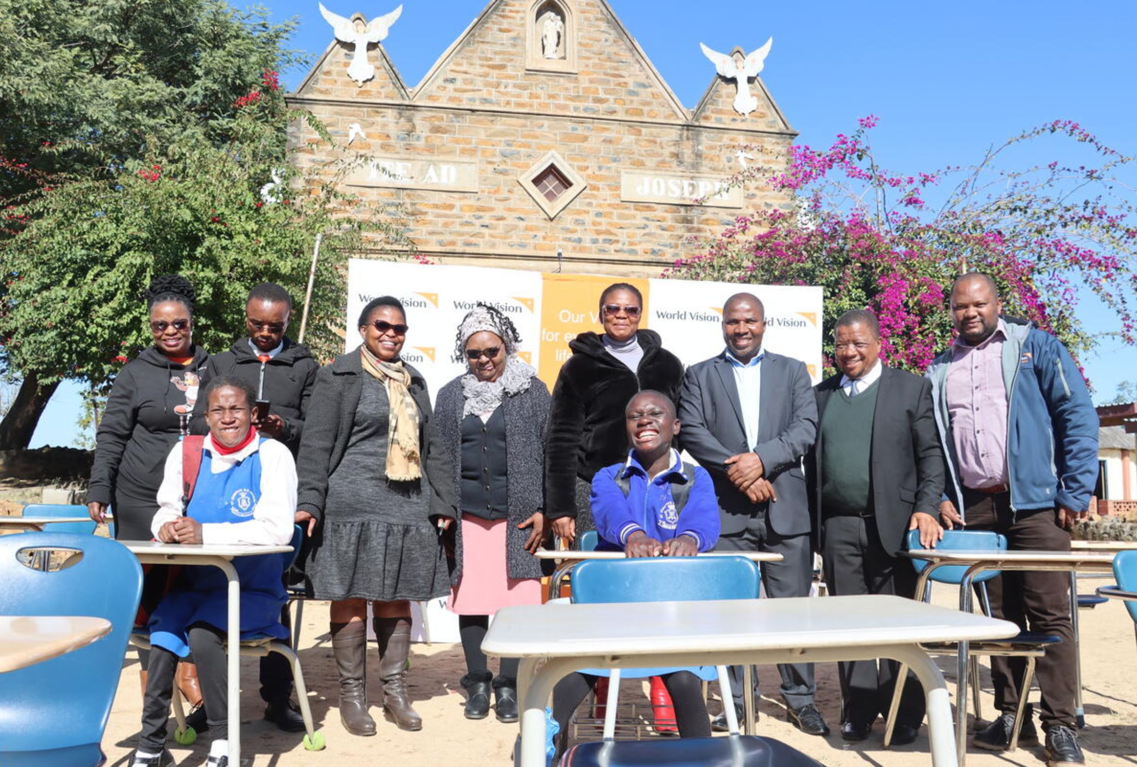 Teachers and two students smile at the camera outside a school while sat at desks