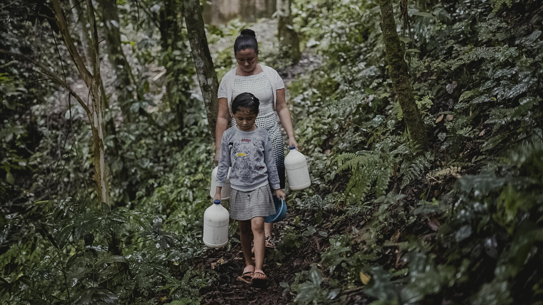 A mother and daughter in Honduras walking through woods both carrying water contrainers in their hands. 