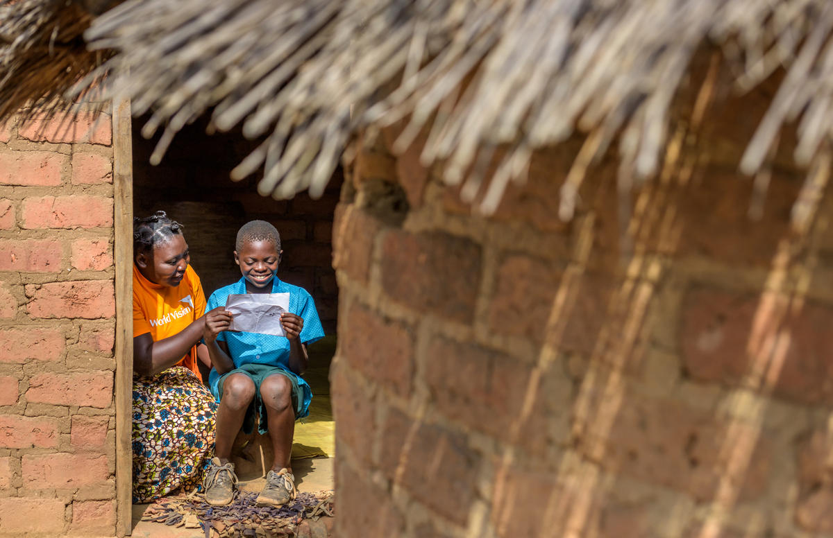 Child in Zambia reads a letter from his sponsor while a World Vision worker sits next to him