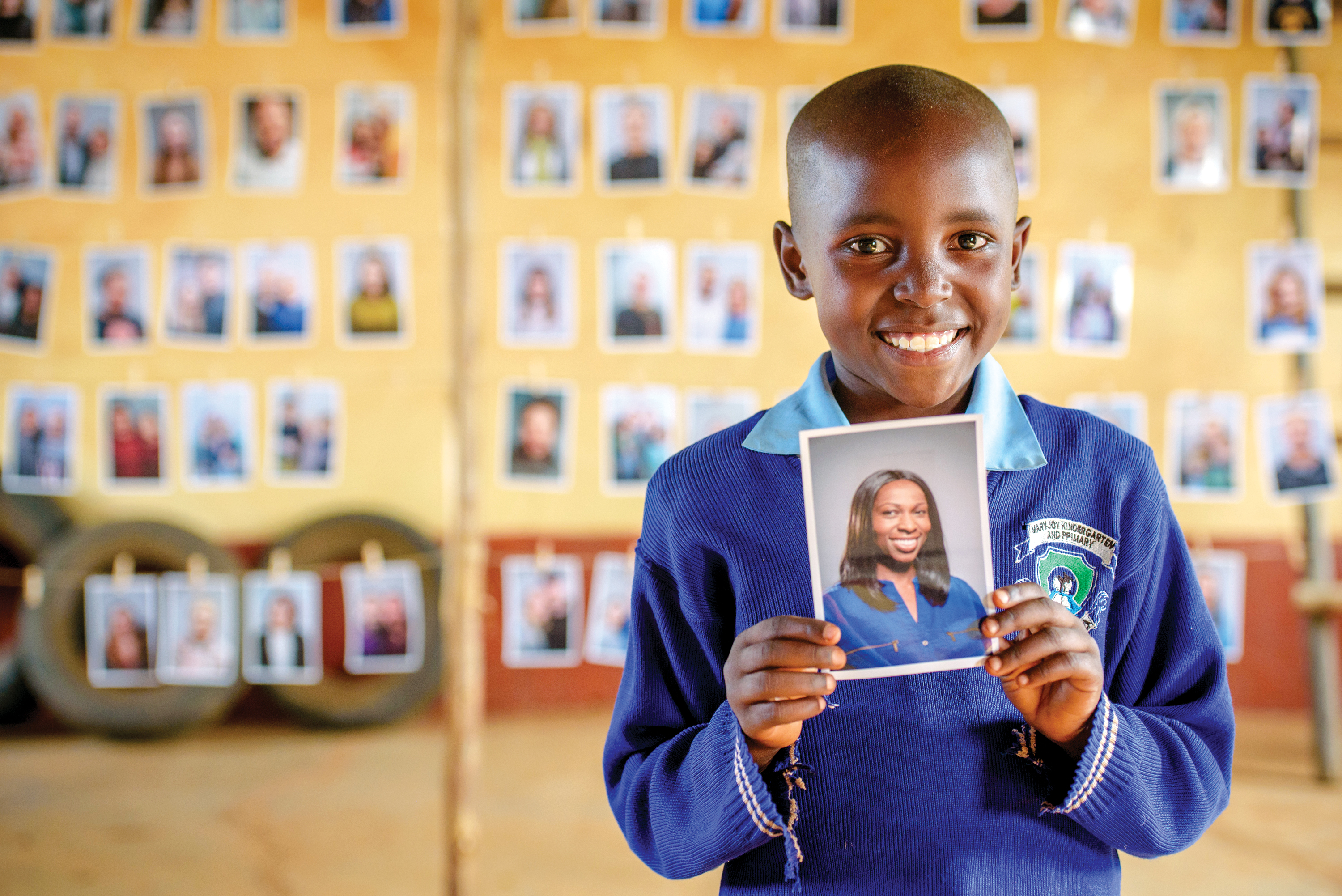 A young girl in Kenya stands in front of rows of photos. She's holding a photo of the sponsor she chose and smiling at the camera.