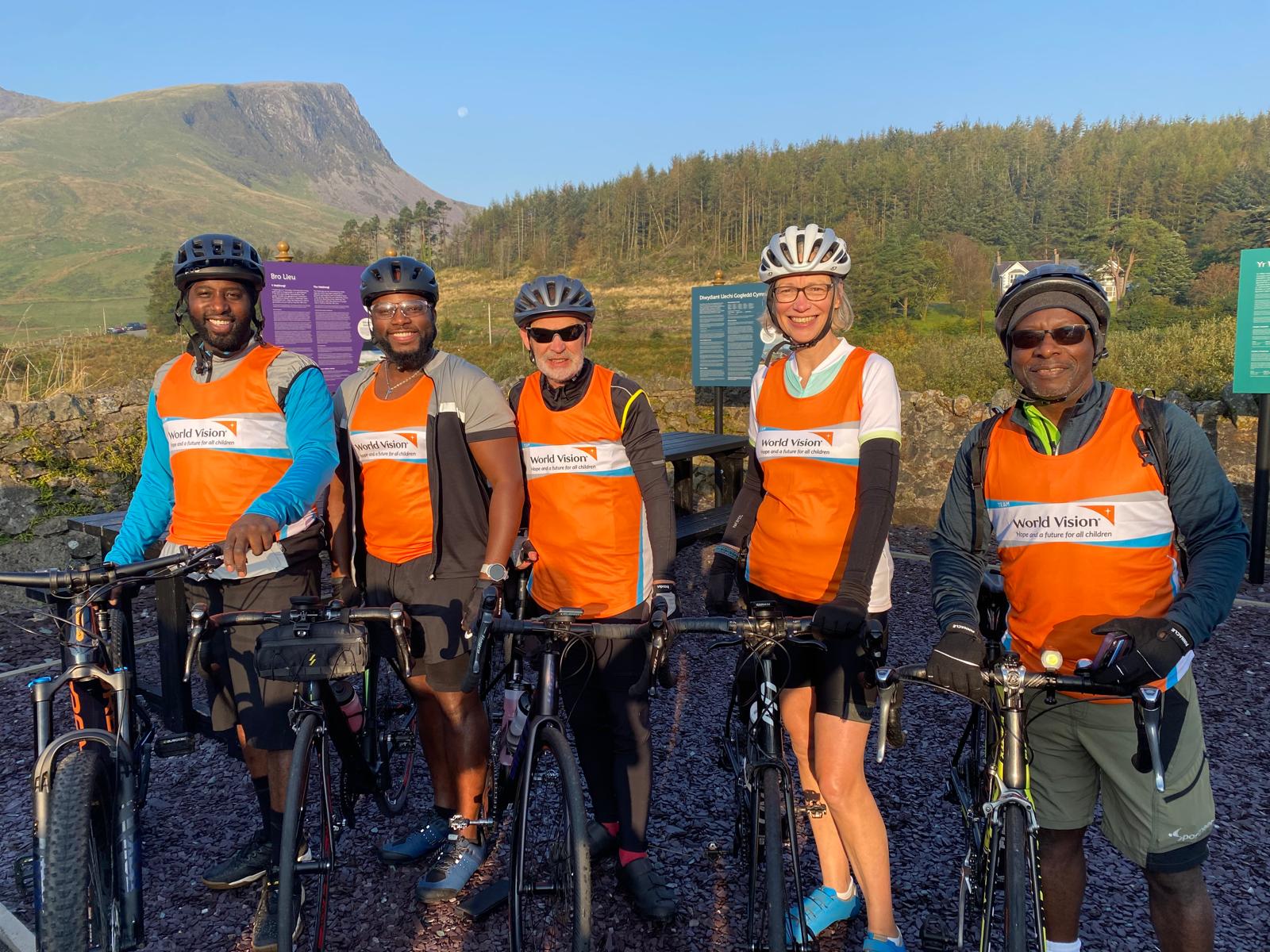 A group of cyclists pose for a photo. They have their helmets and orange World Vision branded vests on.