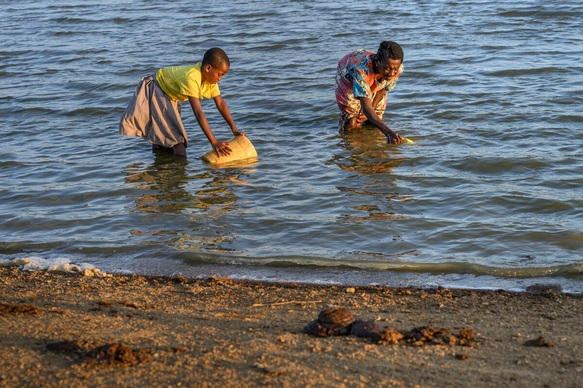 A girl and her grandmother fetch water into their buckets, they are standing in a stream of water