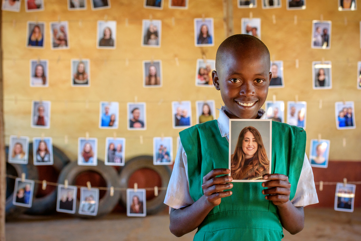 Rosemary from Kenya smiles and holds up a photo of the sponsor she has chosen. There are rows of photos of other sponsors in the background behind her