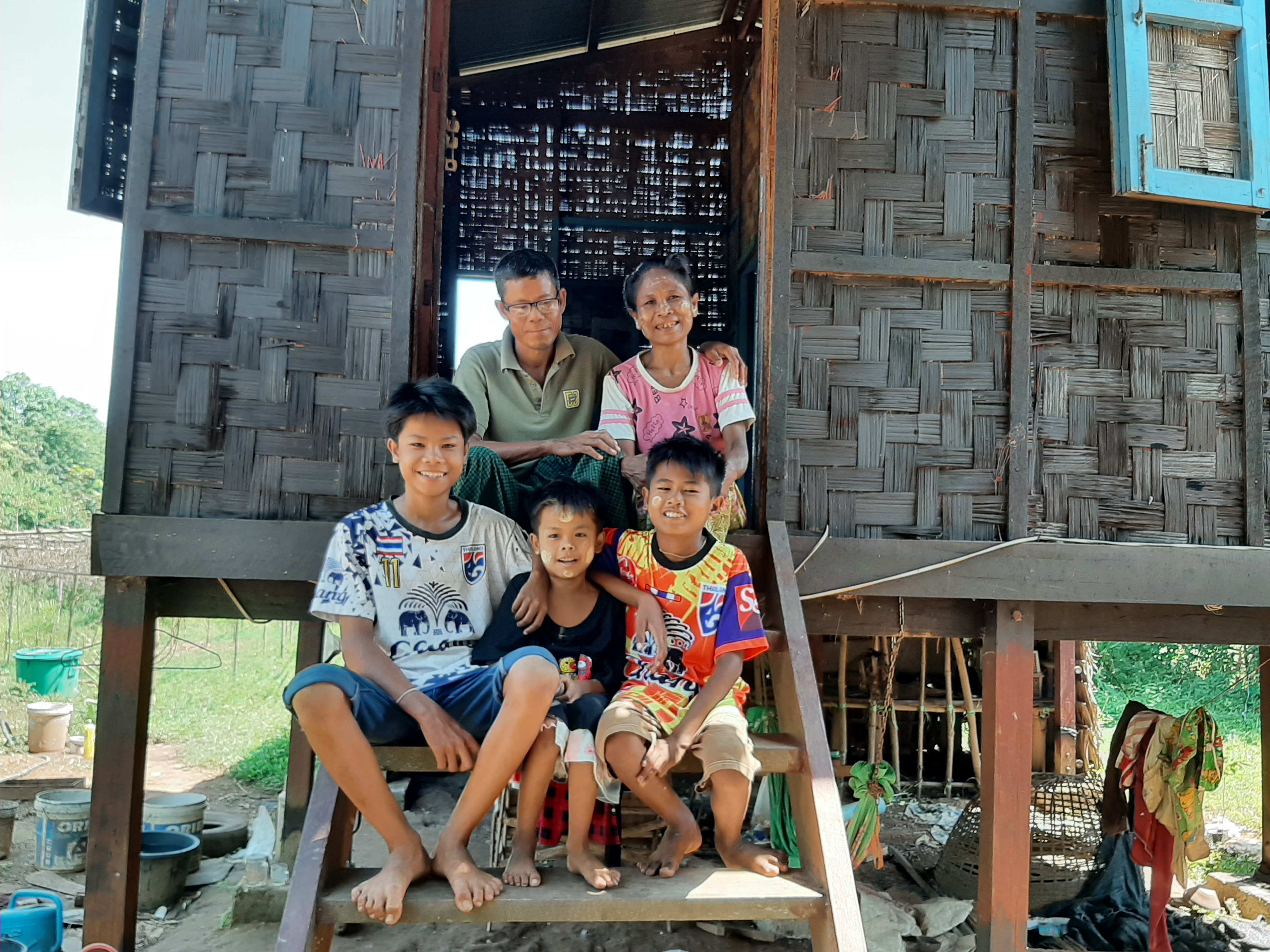 Parents and their three sons from Myanmar sat on wooden steps leading into their wooden hut