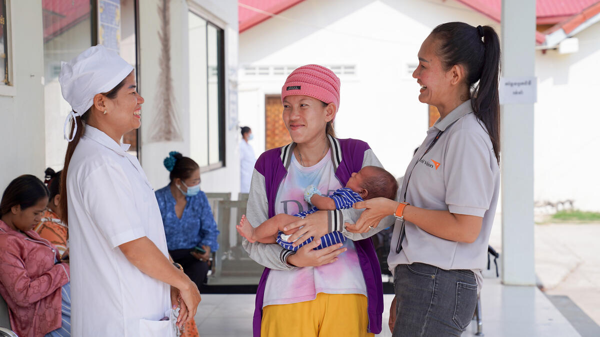 A mum in Cambodia at a health clinic holding her baby and smiling with two heath workers next to her