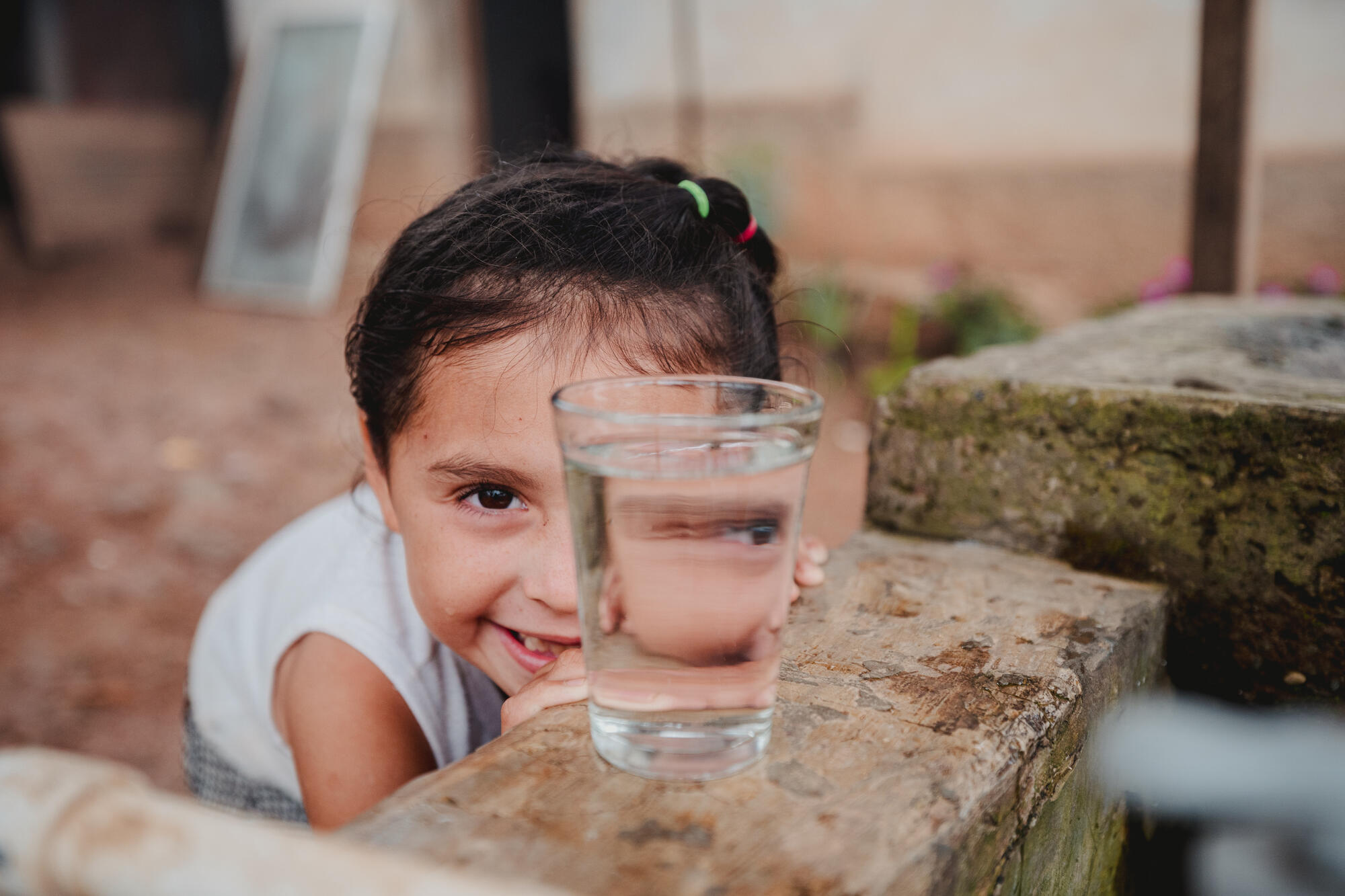 Girl from Honduras smiles while crouching behind a glass of clean water on a wall.