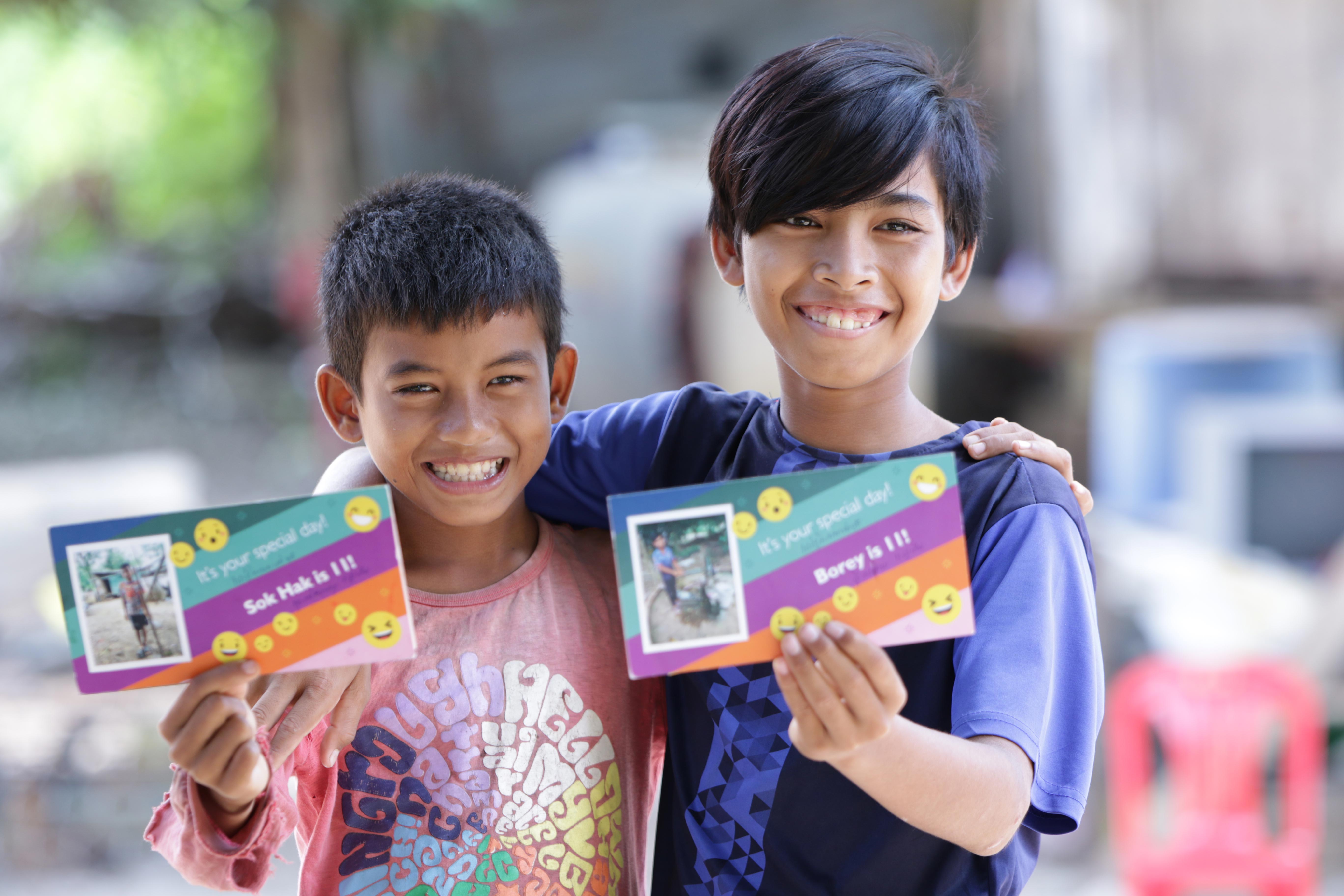 Two Cambodian children smile as they hold up birthday cards from their sponsors