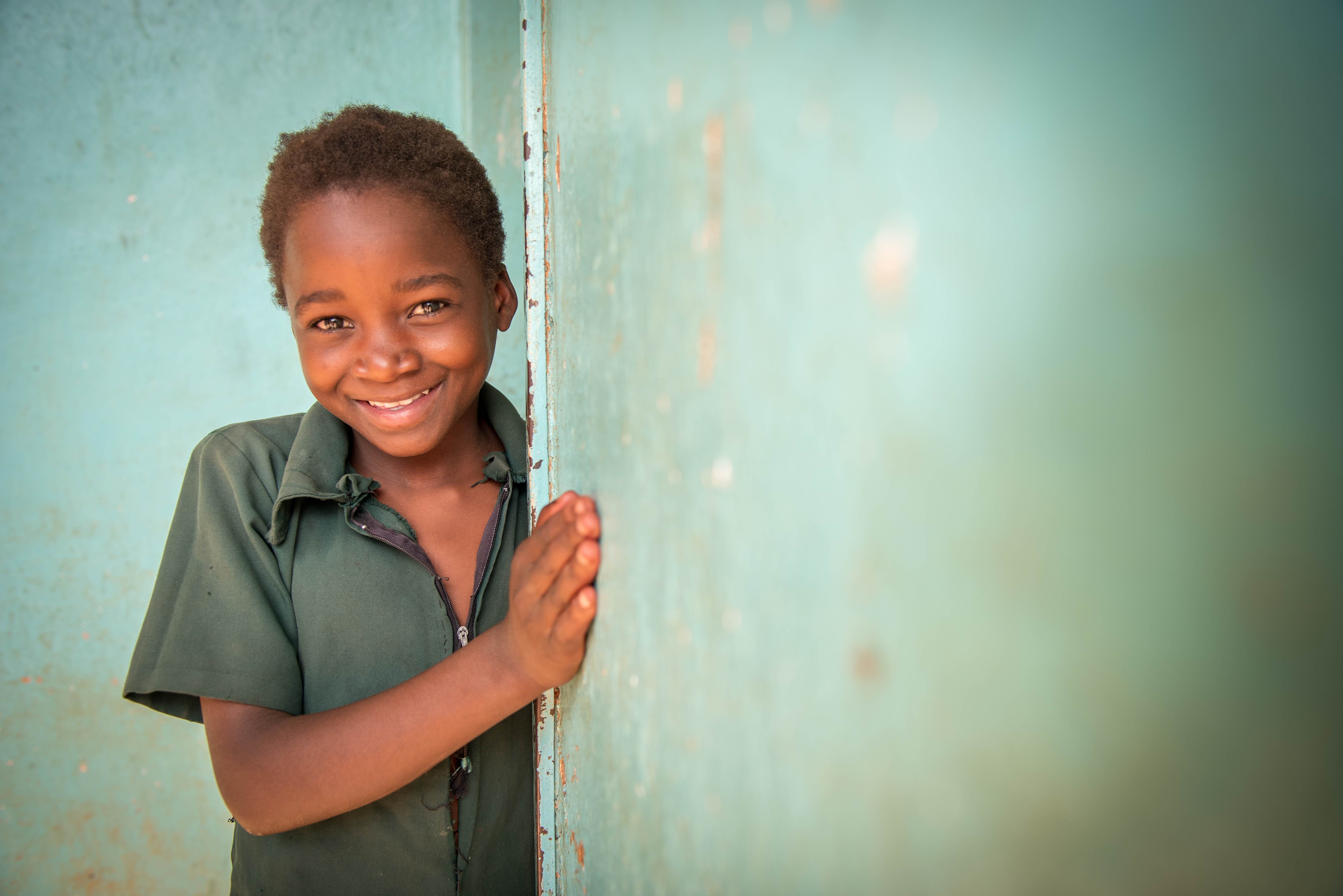 Girl peeks out from the side of a green wall, smiling at the camera, in Zambia