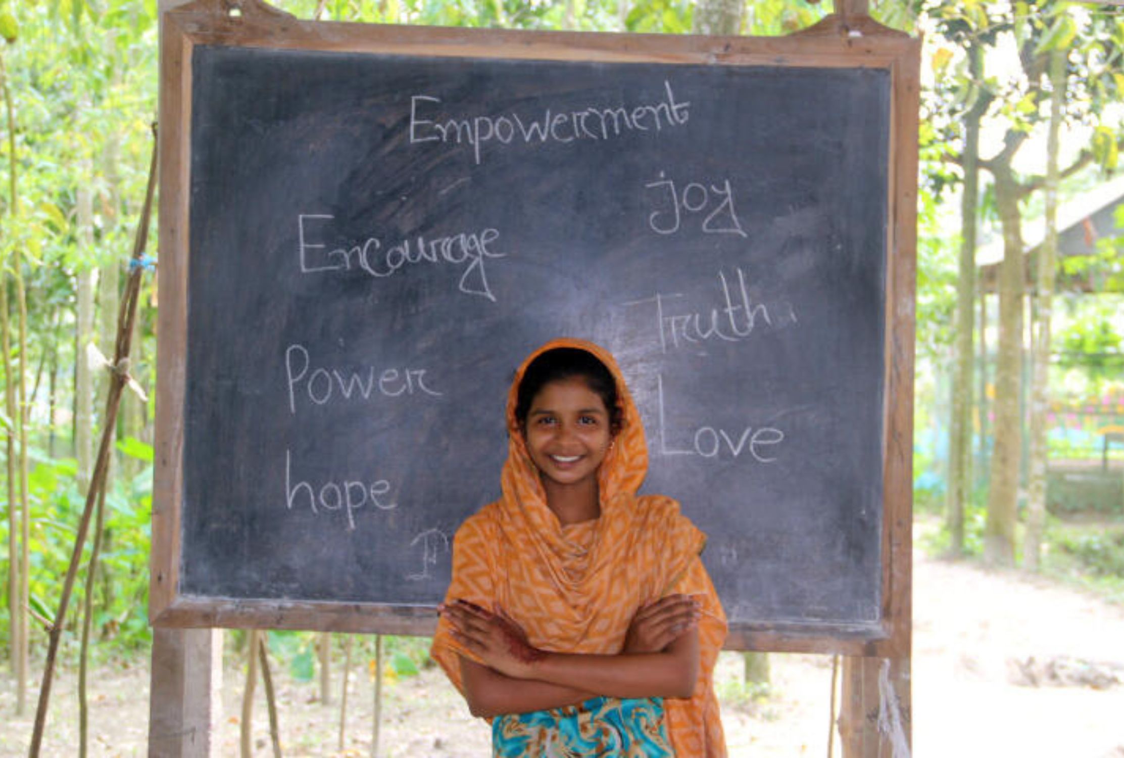 Girl in Bangladesh smiles, standing in front of a blackboard displaying empowering messages